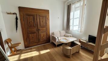 Una habitación con un armario de madera, un sofá, una mesita con papeles, un televisor y una ventana con cortinas.