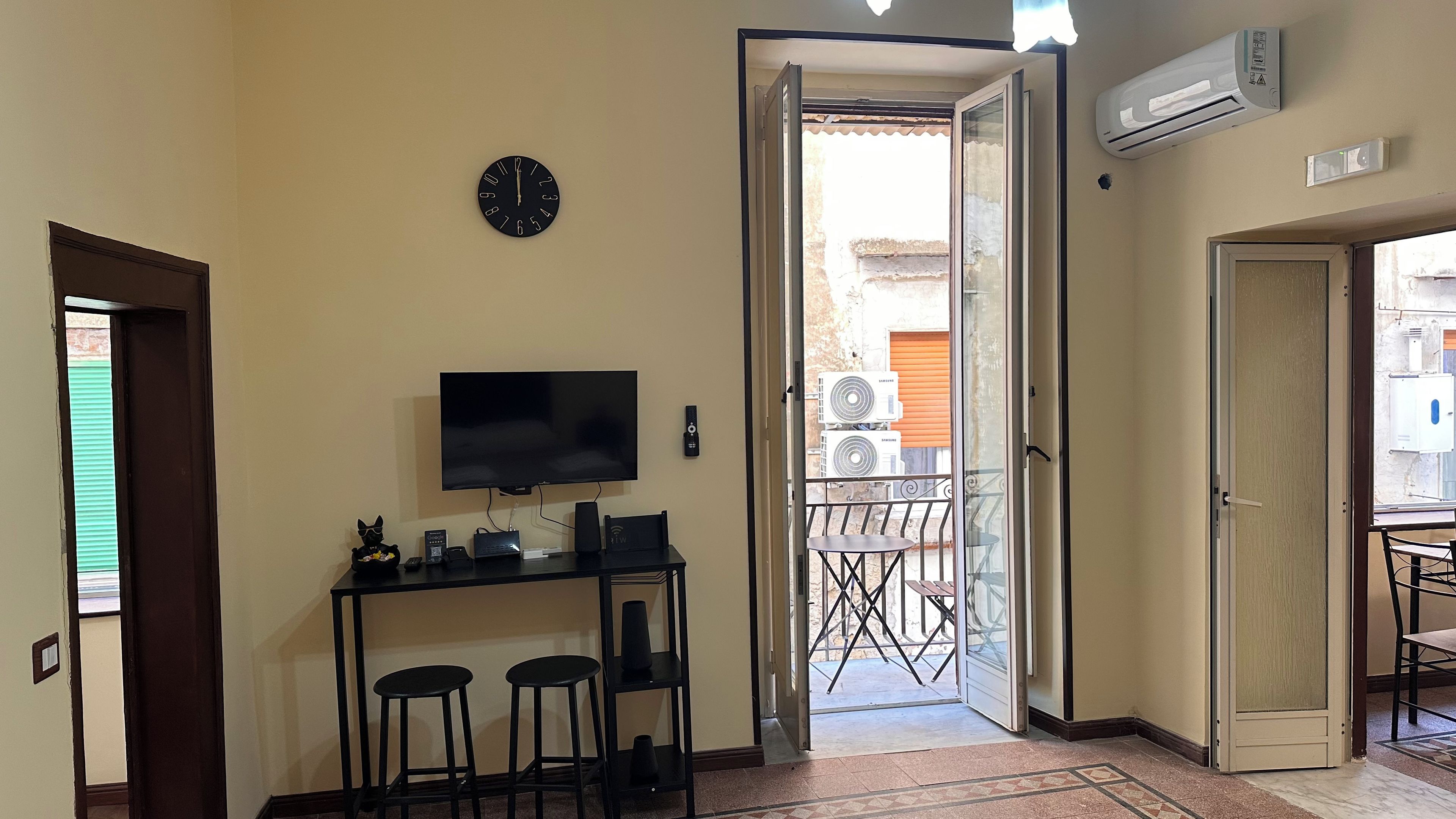 A room with a TV, a clock, and a door leading to a balcony with outdoor furniture.