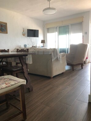 A living room with a sofa, a dining table with chairs, a TV mounted on the wall, and a window with blinds. - New Listing (Cabo de Palos)