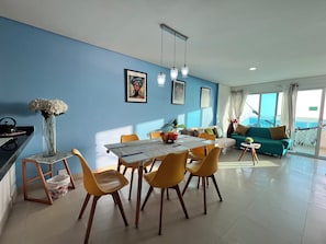 Un soggiorno moderno con zona pranzo, caratterizzato da una parete blu, un divano, un tavolo con sedie e vista sul mare attraverso ampie finestre.