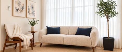 Una sala de estar con un sofá color beige, una silla de madera, una mesita con una planta y un ventanal con cortinas traslúcidas.
