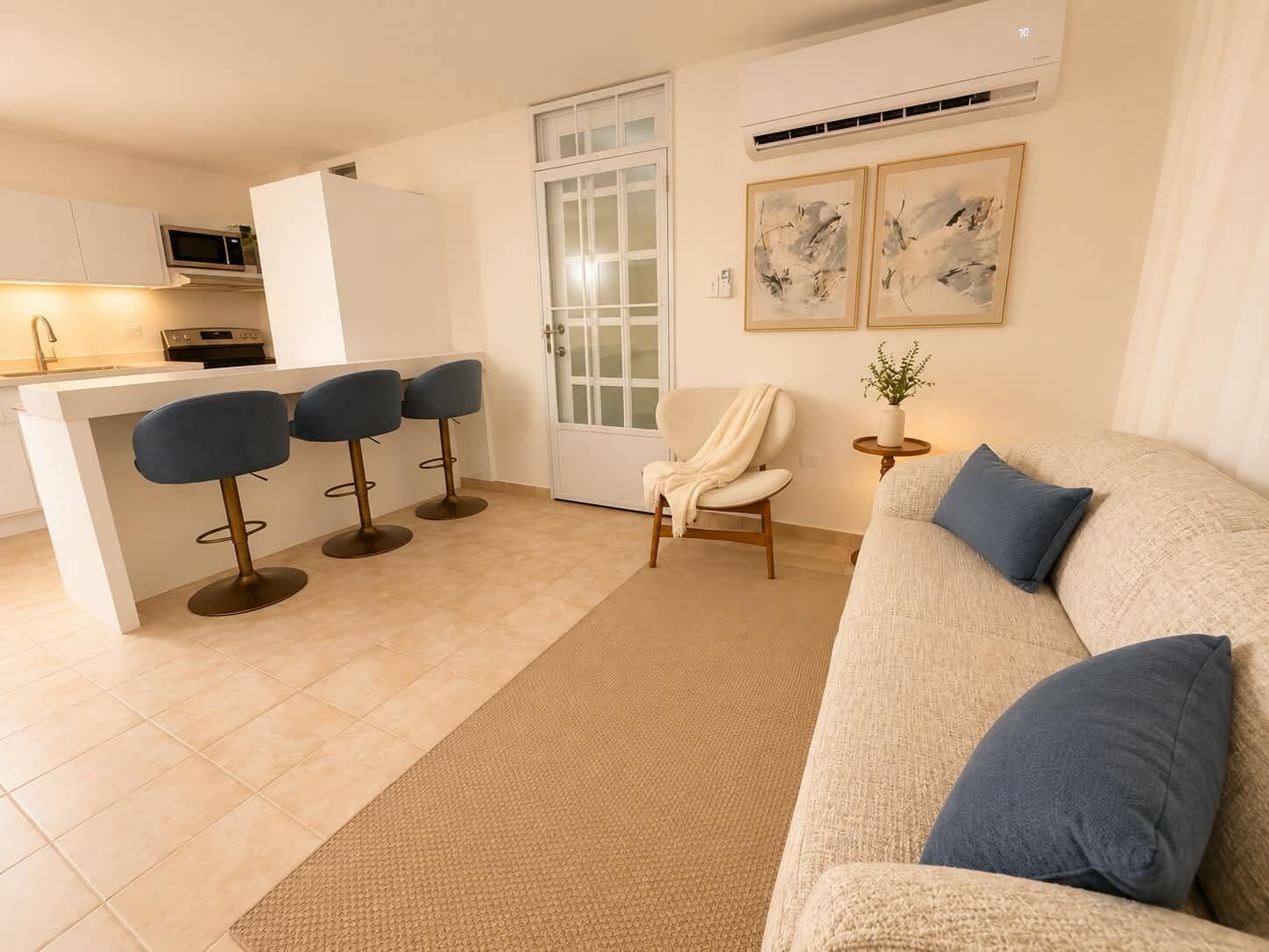 Una sala de estar moderna con un sofá beige, una mesa de centro y una mesita auxiliar con una planta. Hay una zona de cocina con un microondas y un taburete de bar.