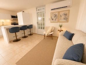 Una sala de estar moderna con un sofá beige, una mesa de centro y una mesita auxiliar con una planta. Hay una zona de cocina con un microondas y un taburete de bar.
