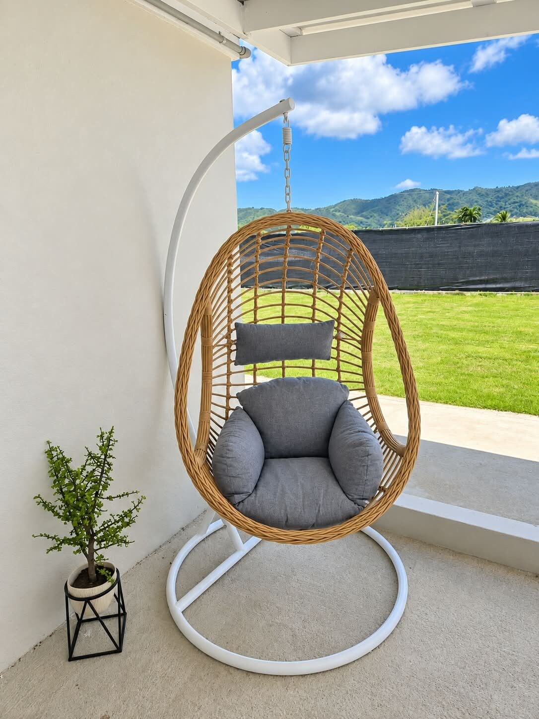 A wicker hanging chair with grey cushions on a balcony with a view of a green landscape and hills.