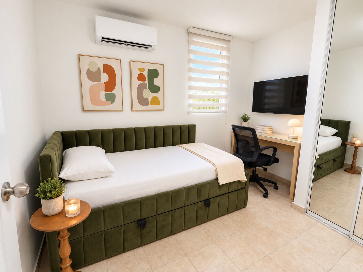 A bedroom with a green upholstered bed, a desk with a chair, a television, and a small table with a plant and candle.