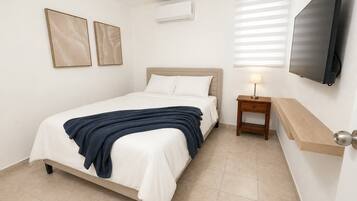 Habitación de hotel con cama, mesita de noche con lámpara, televisor montado en la pared y dos cuadros enmarcados.