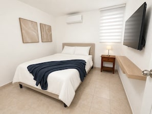 Habitación de hotel con cama, mesita de noche con lámpara, televisor montado en la pared y dos cuadros enmarcados.