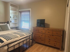 Ein Schlafzimmer mit einem Bett, einer Holzkommode, einem Fernseher auf der Kommode und einem Fenster mit Jalousien.