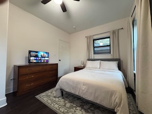 Une chambre à coucher avec un lit, une commode, un téléviseur posé sur la commode et une fenêtre avec des rideaux.