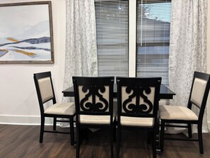 Un coin repas avec une table et des chaises, une fenêtre avec des stores et un tableau accroché au mur.