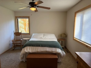 Ein Schlafzimmer mit einem Bett, einem Sessel, einem kleinen Tisch, einem Deckenventilator und einem Fenster mit Jalousien.