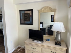 A hotel room with a television on a dresser, a lamp, a framed picture, and a mirror. - Cozy 1-bedroom condo in pleasant Daytona Beach with AC and gym (Daytona Beach)