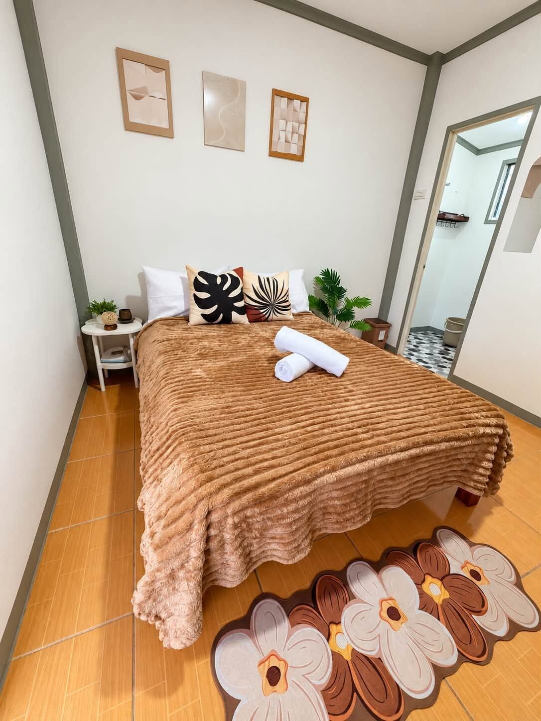 A neatly made bed with a brown blanket, pillows with floral patterns, and a rolled white towel. There are two framed artworks on the wall above the bed.