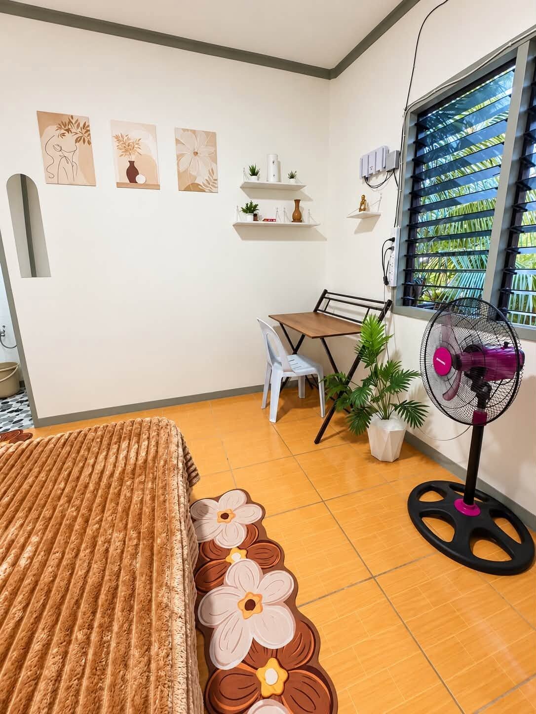 Et værelse med seng, skrivebord, stol, en potteplante, en ventilator og billedkunst på væggen.
