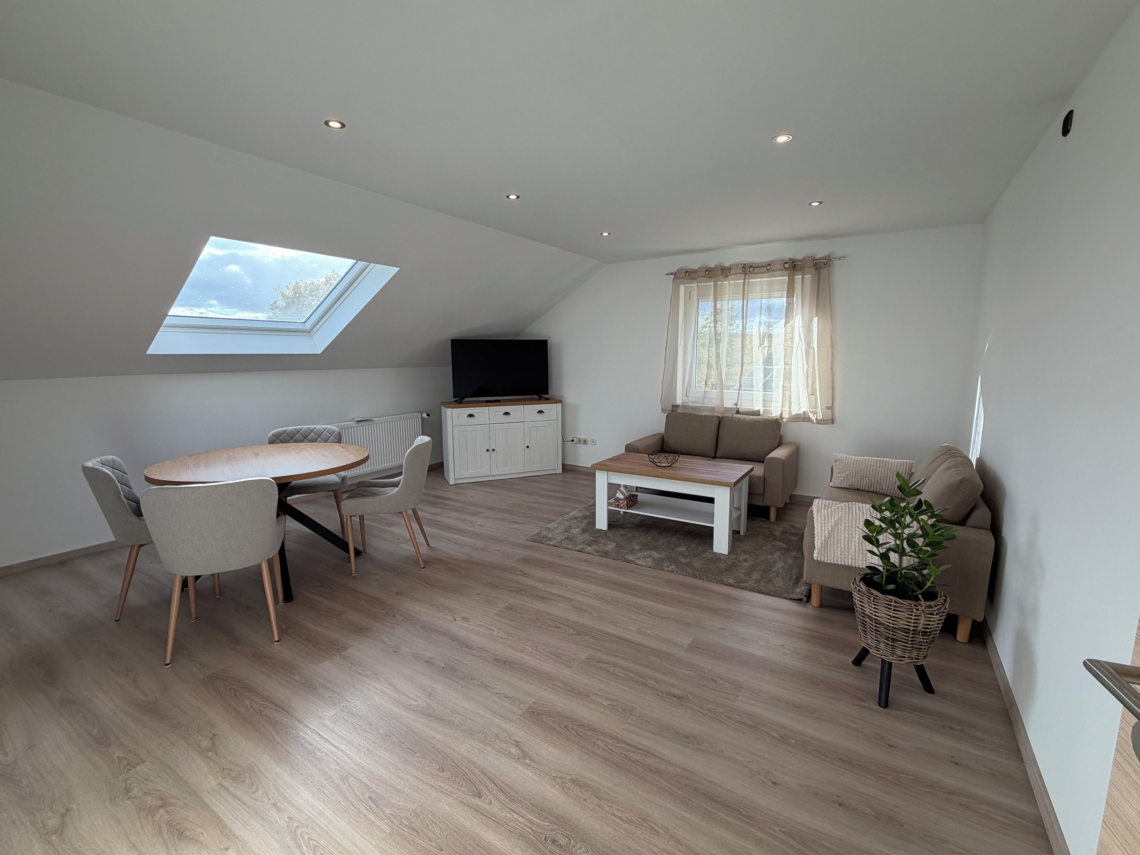 A modern living room with a dining area, a sofa, a TV, and a skylight.