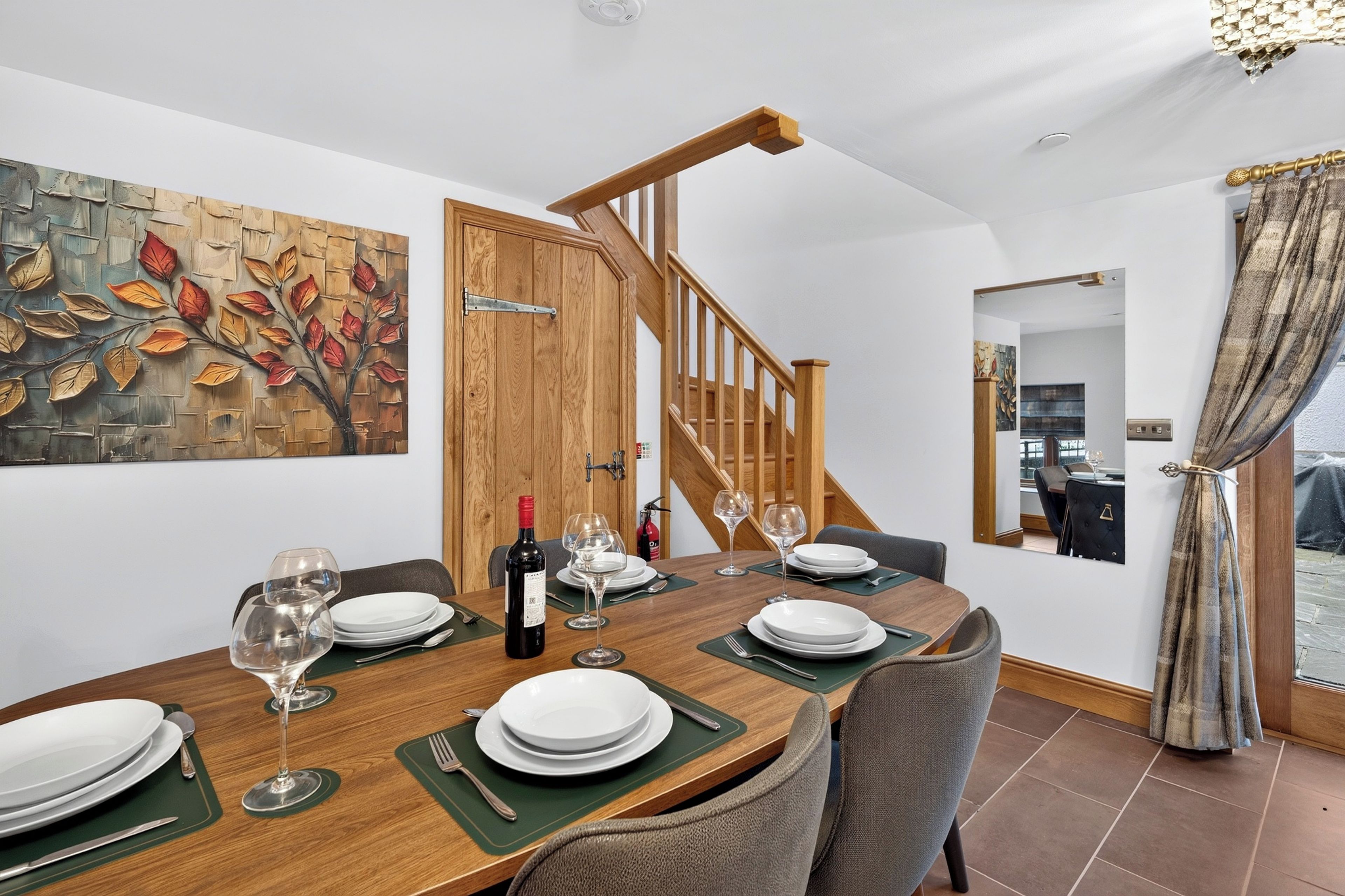 A dining area with a wooden table set for four, a bottle of wine, and a painting of leaves on the wall.