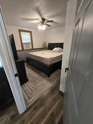 A bedroom with a bed, a ceiling fan, a window with blinds, a lamp, and a television. - Cozy 1-bedroom apartment in enchanting Sparta surounded by trees and wilderness. (Sparta)