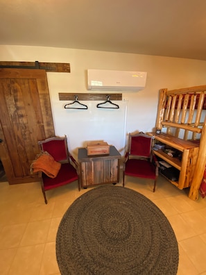 Una habitación con puerta de madera, dos sillas rojas, una mesita con una caja etiquetada 'WINCHESTER', un tapete redondo y un estante de madera.