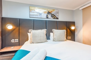Hotellihuone, jossa on suuri sänky, kaksi yöpöytävalaisinta, seinälle ripustettu taideteos ja puinen yöpöytä, jossa on kaukosäädin.