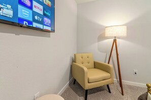 Een kamer met een televisie waarop verschillende streamingdiensten worden getoond, een beige fauteuil, een vloerlamp en een vloerkleed.