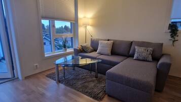 Een moderne woonkamer met een hoekbank, een glazen salontafel en een groot raam met zonwering.