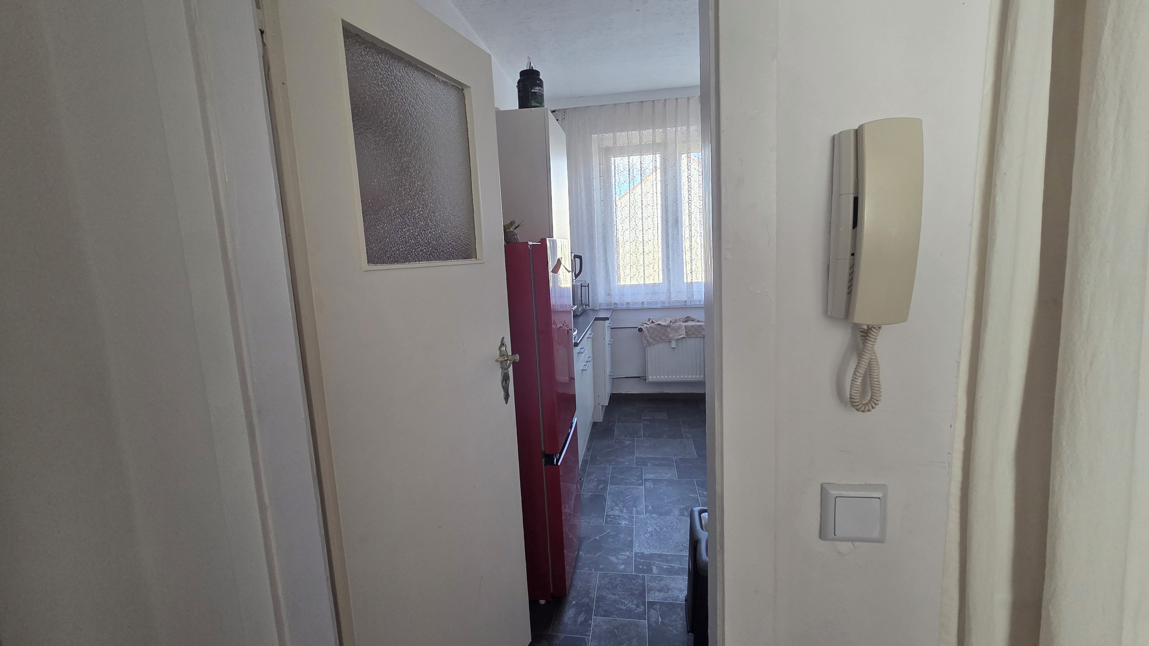 Ein schmaler Flur mit einem roten Kühlschrank, einem Telefon und einem Fenster mit Vorhängen.