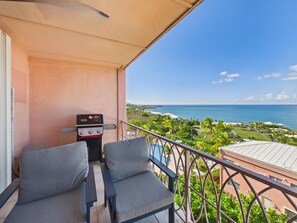 Un balcon avec deux chaises grises, un barbecue et une vue sur l’océan et la zone touristique.