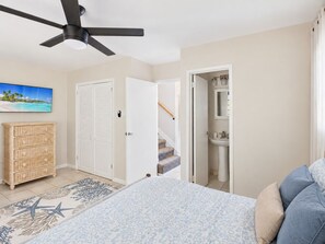 Une chambre à coucher avec un lit, un ventilateur de plafond, une commode en bois et une salle de bain avec un lavabo, ainsi qu’un escalier.