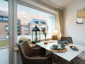 A dining area with a table set for two, a window with a view of buildings, and a lamp on the table. - Holiday Apartment With 1 Bedroom (Sylt)