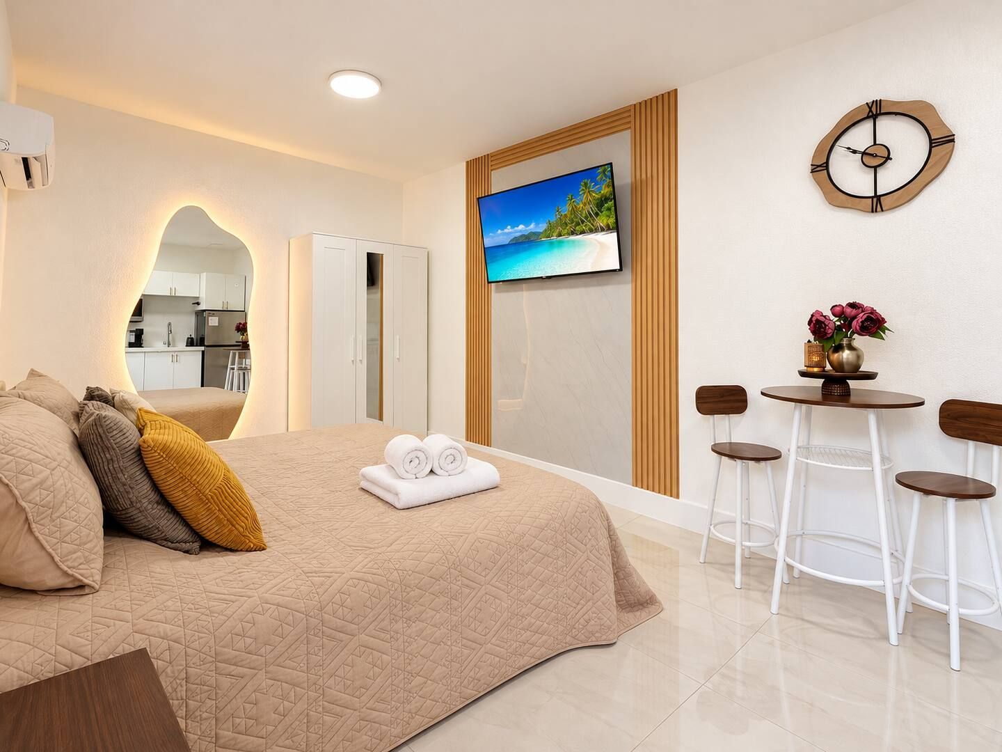 A hotel room with a bed, a TV, a small table with chairs, and a wall-mounted clock.