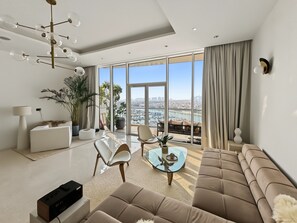 A modern living room with a sofa, chairs, a coffee table, and a large window offering a city view. - Tiara Aquamarine - 705 3BD (Dubai)