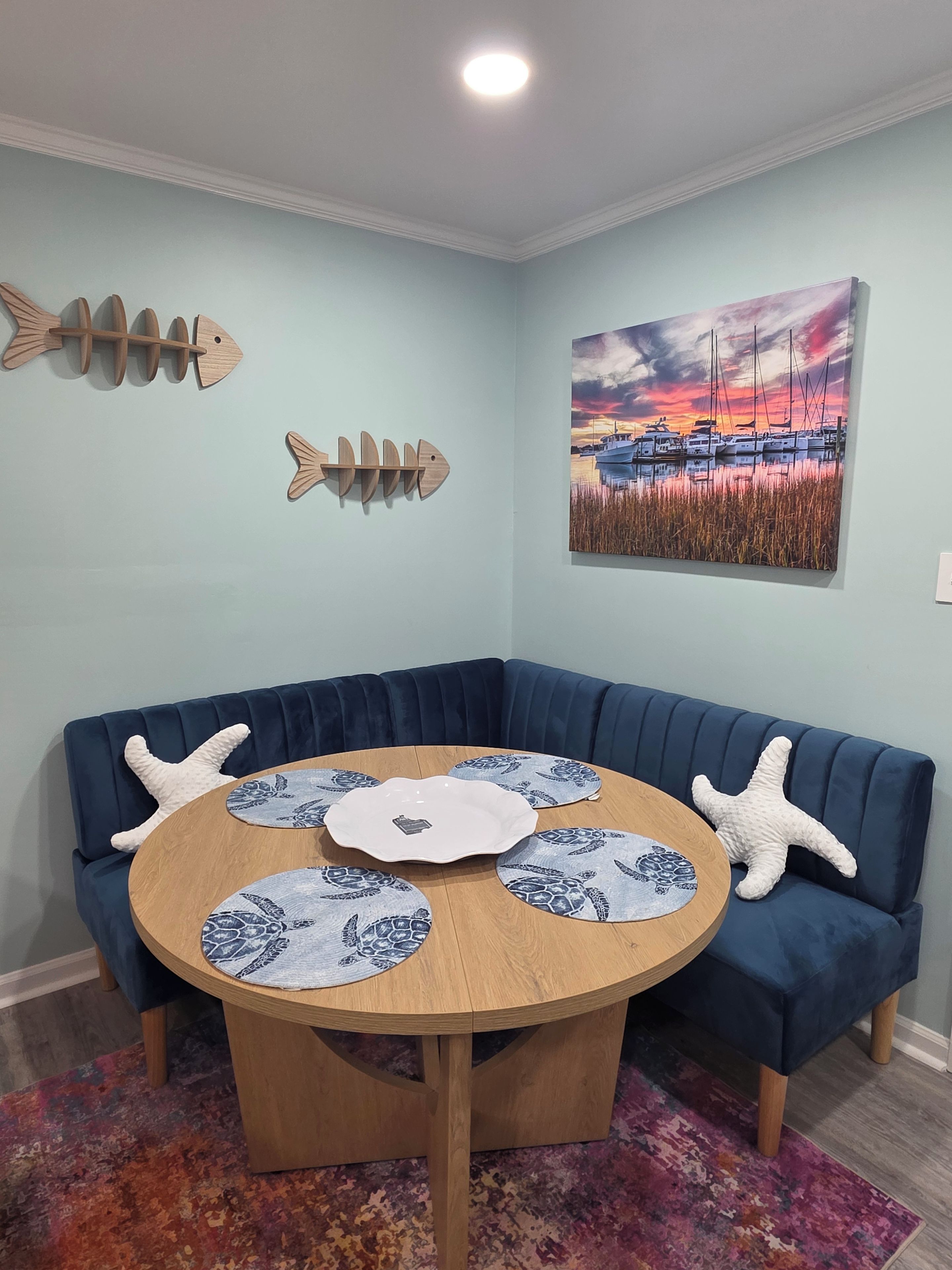 A corner room with a blue upholstered bench, a round wooden table with turtle placemats, starfish pillows, and a painting of a sunset over boats.
