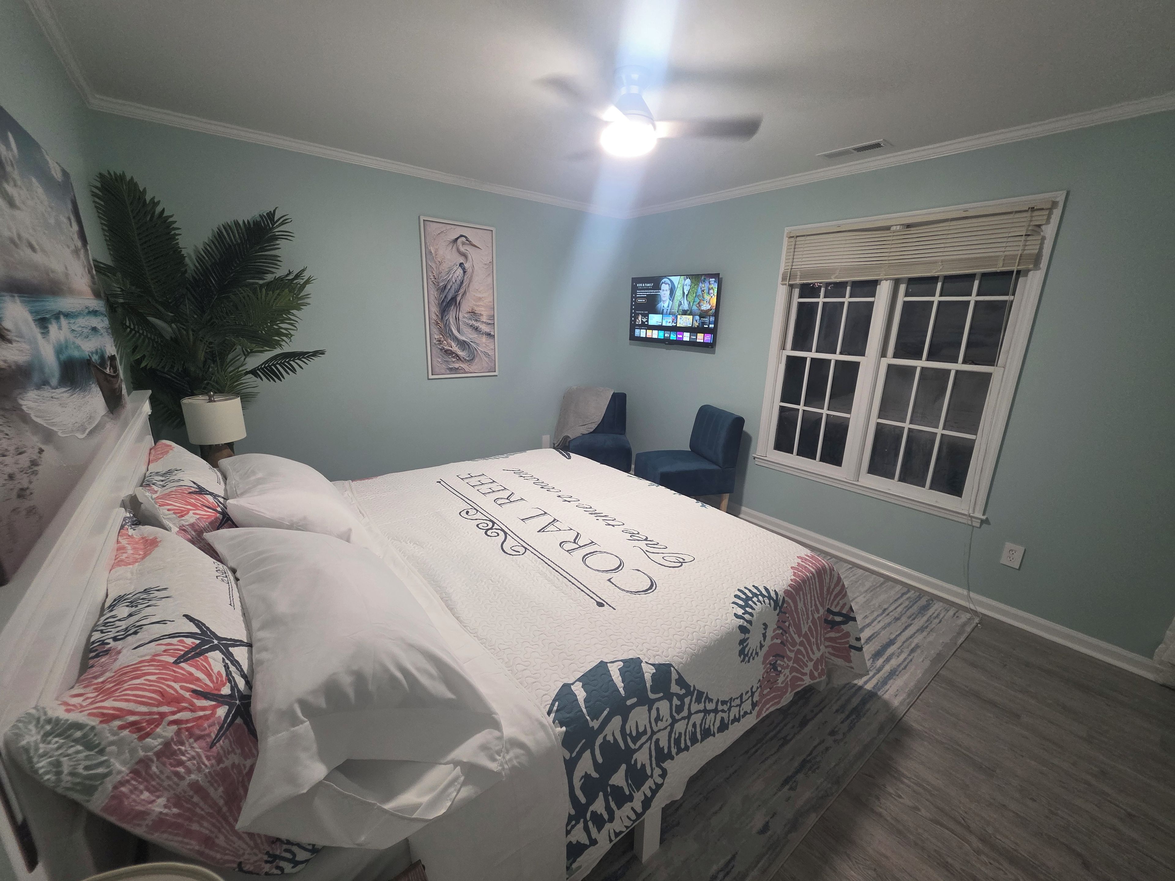 Une chambre à coucher avec un lit, un ventilateur de plafond, une télévision, une fenêtre et une plante.