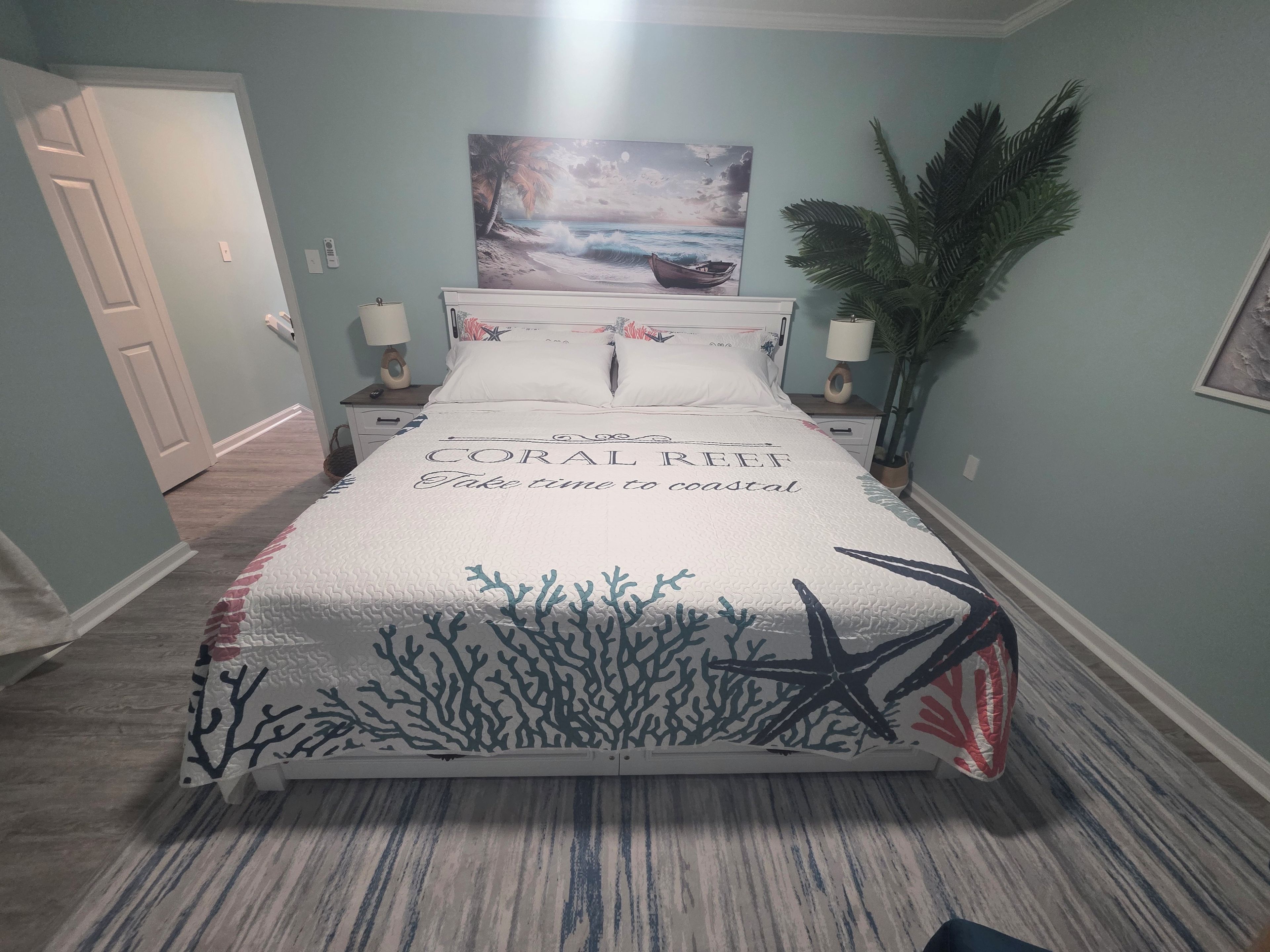 A bedroom with a bed featuring a coral reef design, a painting of a beach scene, and a potted plant.