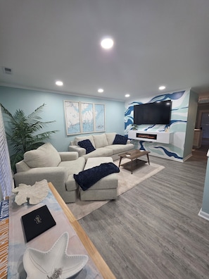 A modern living room with a sectional sofa, a flat-screen TV, and a decorative wall with a wave pattern. - 2-bedroom condo in enchanting Morehead City minutes away from Atlantic Beach (Morehead City)