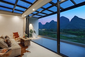 A modern living room with a large glass wall, a sofa, a wooden coffee table, and a potted plant.
