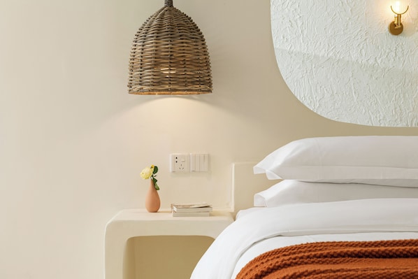 A bedroom with a bed, a bedside table with a vase and a book, a wall-mounted light fixture, and a textured wall.