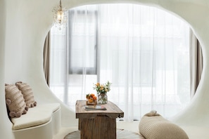 A room with a round window, a sofa, a wooden table with a vase of flowers, and a hanging light fixture.