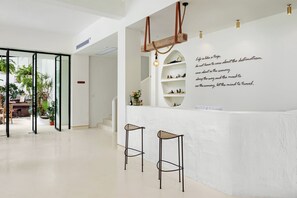 A modern interior with a reception desk, two bar stools, a wall-mounted shelf with plants, and a ceiling light fixture.
