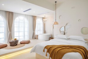 Ein modernes Schlafzimmer mit einem großen Bett, einem runden Spiegel, einer Chandelüster und Blick ins Freie.