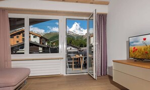 Une pièce dotée d’une porte-fenêtre coulissante donnant sur un balcon, d’un téléviseur posé sur un meuble en bois et d’un fauteuil rose.