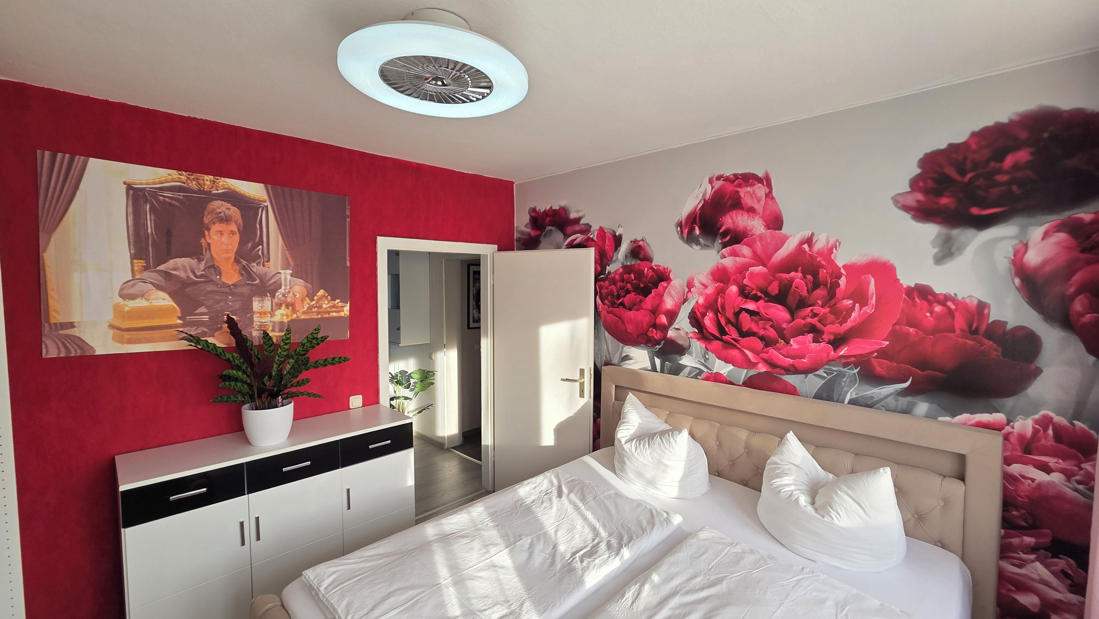 A bedroom with a bed, a dresser, a potted plant, a painting, and a floral wall mural.