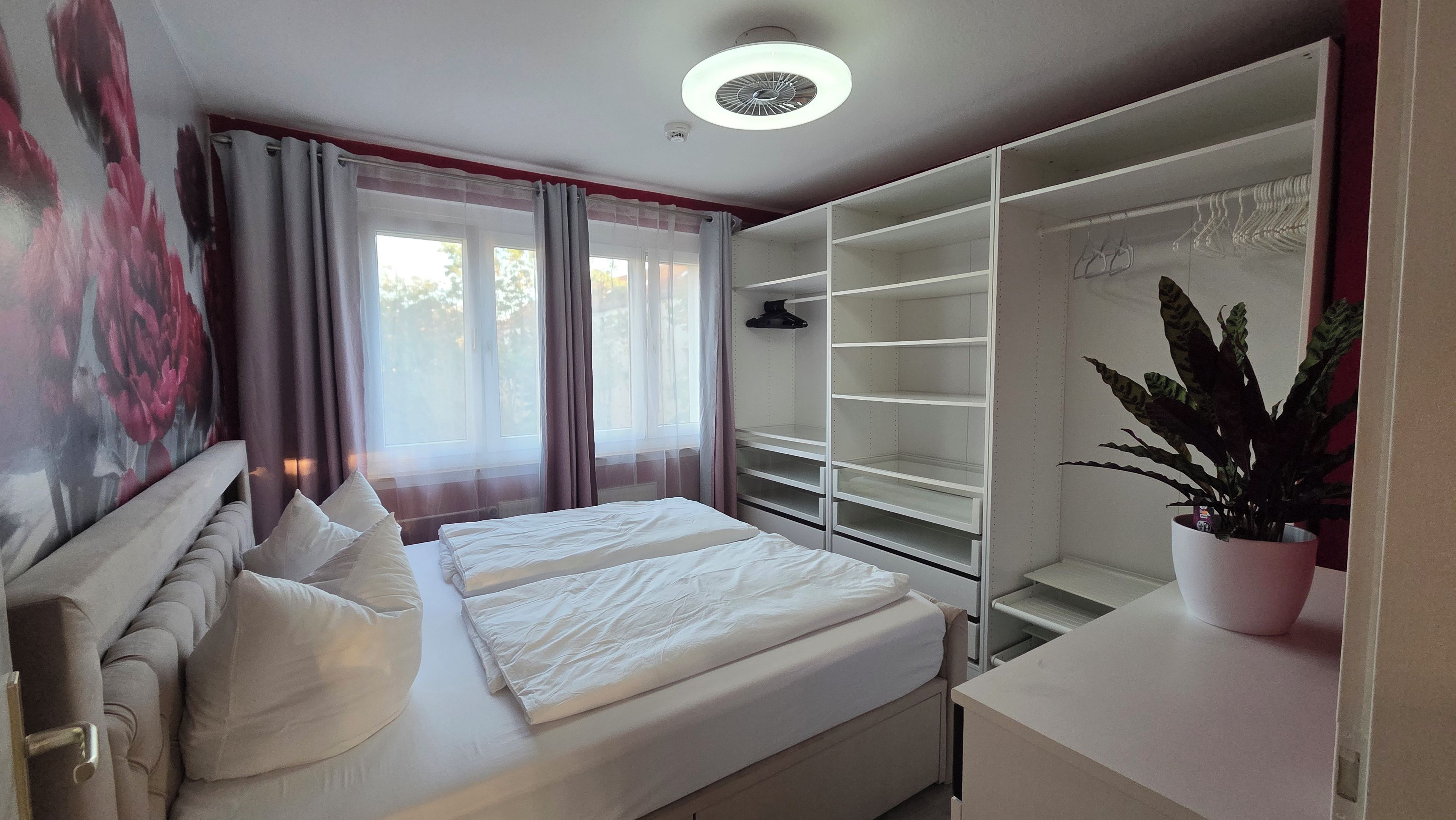 Ein Schlafzimmer mit einem Bett, einem Kleiderschrank, einer Zimmerpflanze und einem Fenster mit Vorhängen.