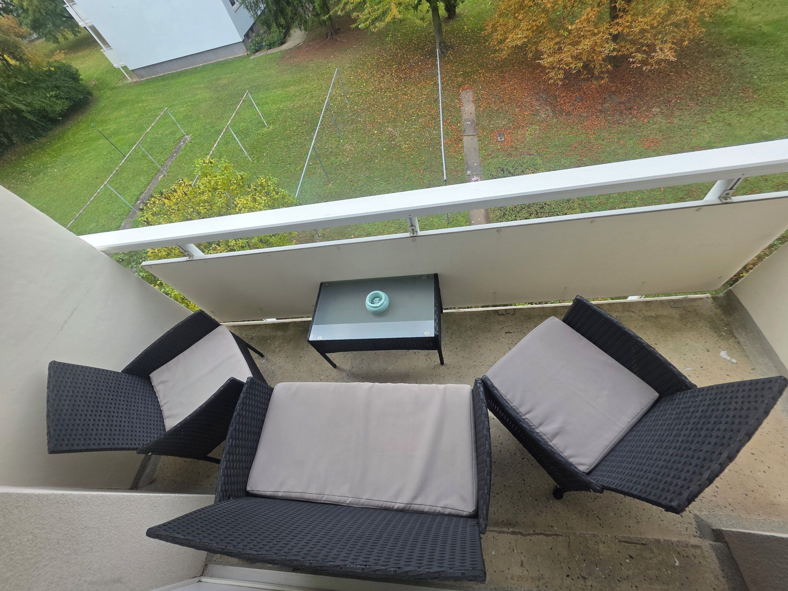 A balcony with two chairs and a small table, overlooking a grassy area with trees and a building.