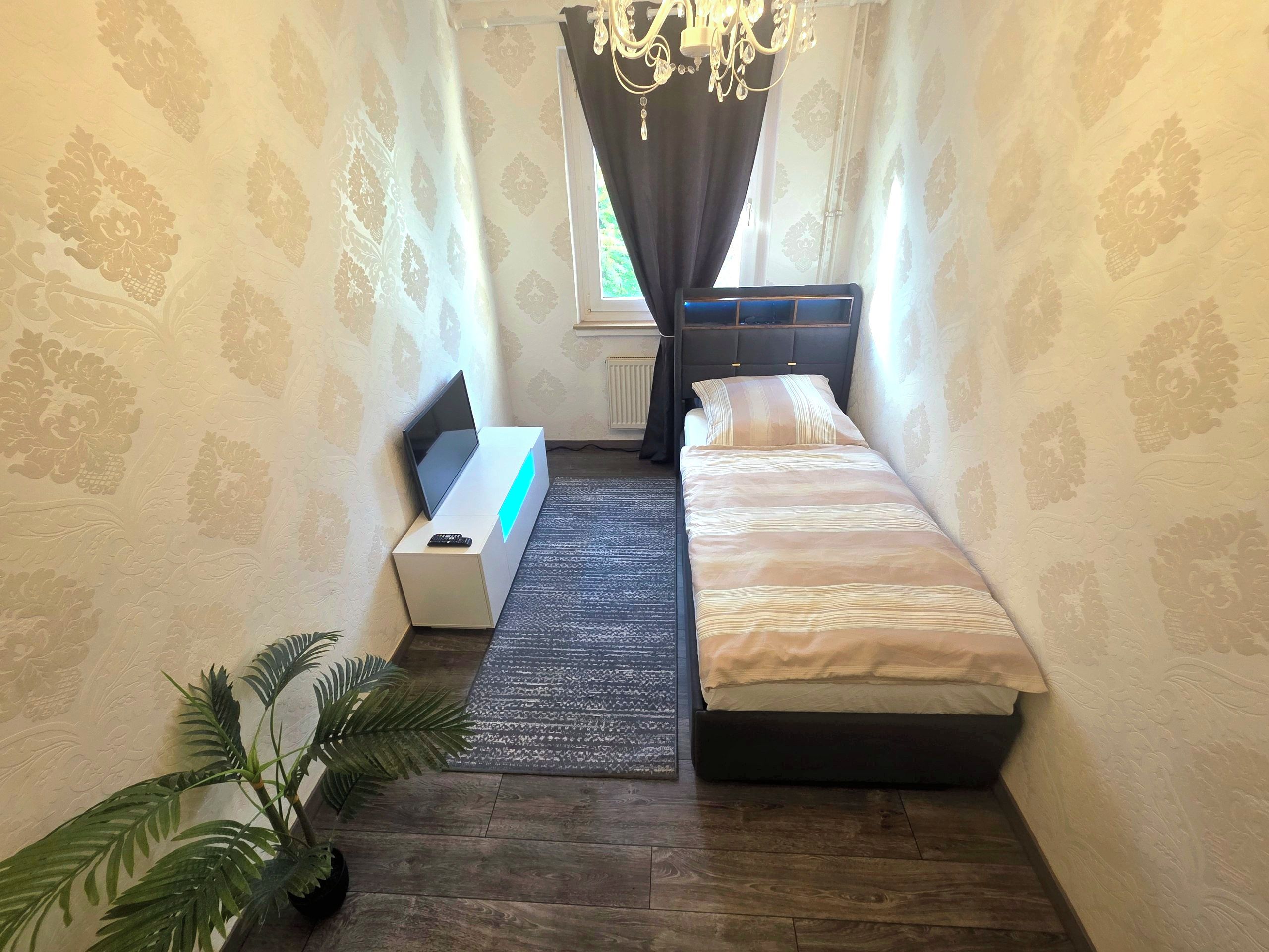 Ein kleiner, schmaler Raum mit einem Bett, einem Schreibtisch mit Computer, einer Zimmerpflanze und einem Fenster mit Vorhängen.