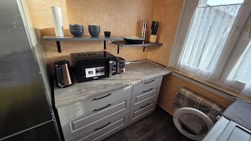 Eine kompakte Küche mit Toaster, Wasserkocher und Mikrowelle. Ein Fenster mit Vorhang und eine Waschmaschine sind ebenfalls vorhanden.