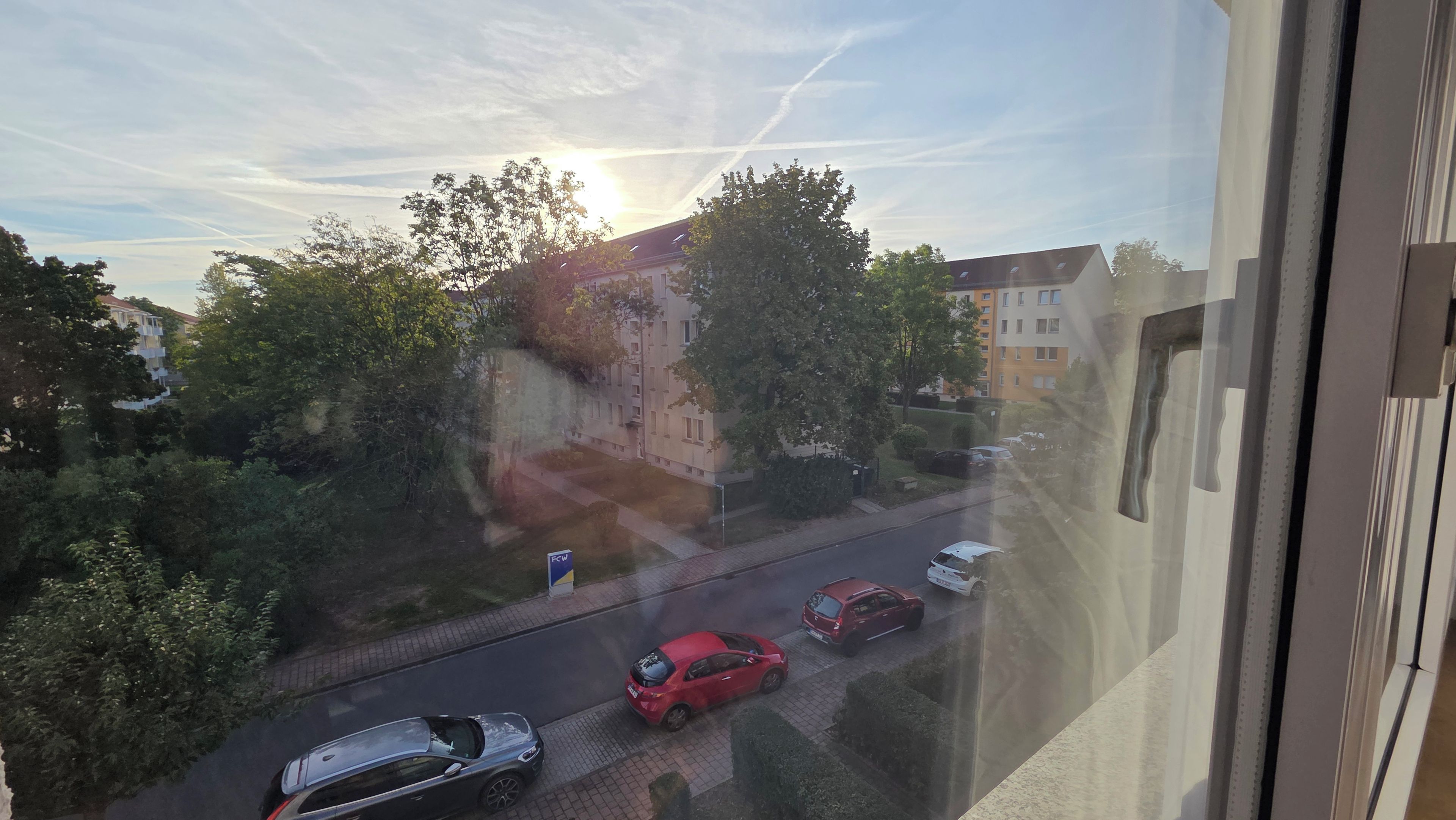 Blick aus einem Fenster auf eine Straße mit parkenden Autos und Gebäuden im Hintergrund.