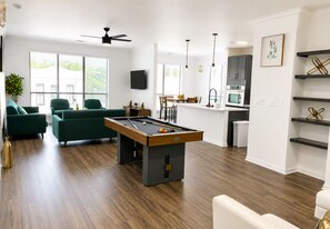 Una sala de estar moderna con mesa de billar, sofás verdes y una zona de cocina con microondas y armarios.