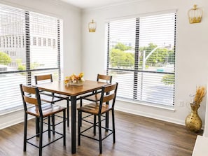 Un comedor con una mesa y sillas de madera, ventanas amplias con persianas y un jarrón con plantas secas en el suelo.