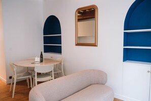 Una habitación con una mesa redonda, sillas blancas, un sofá color beige, un espejo y estanterías azules.